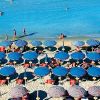 Spiaggia con numerosi ombrelloni blu e persone che camminano sul bagnasciuga del mare azzurro.