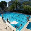 Piscina affollata con persone che nuotano, circondata da palme e edifici residenziali.