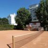 Campo da tennis all'aperto con rete, contornato da alberi e edifici bianchi.